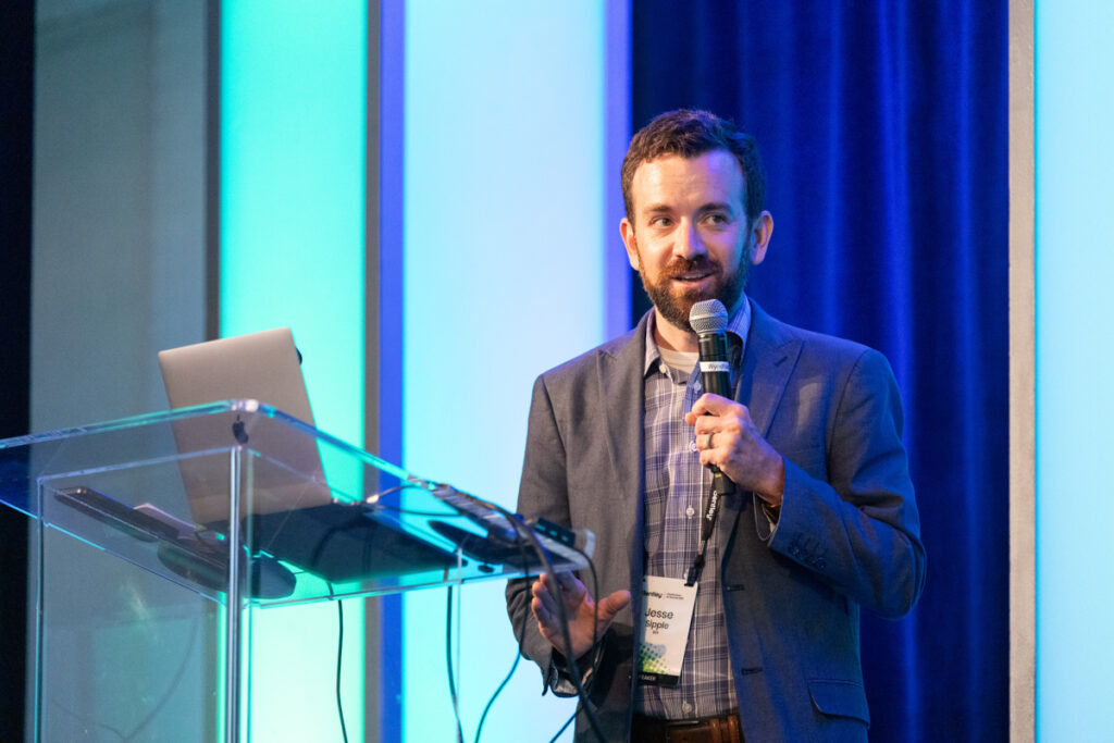 A man with a beard speaks at a podium with a microphone, holding a remote, at a conference with a laptop and a presentation screen behind him.