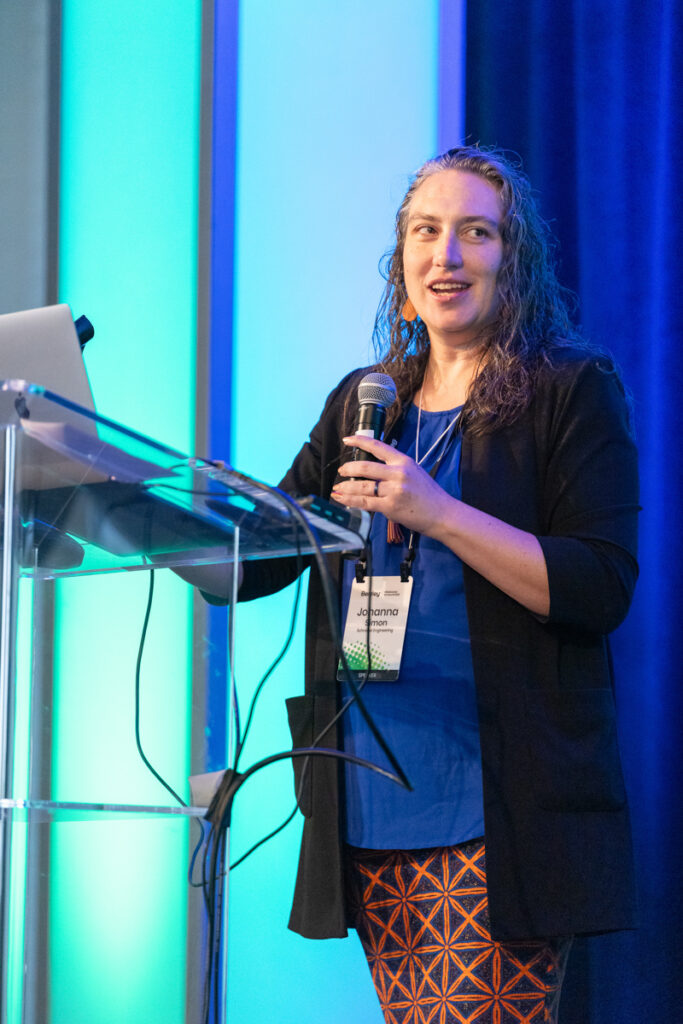 A woman with curly hair stands at a podium with a microphone in a conference room, a name tag reading "joanna" visible. she smiles while speaking, with blue lit panels in the background.