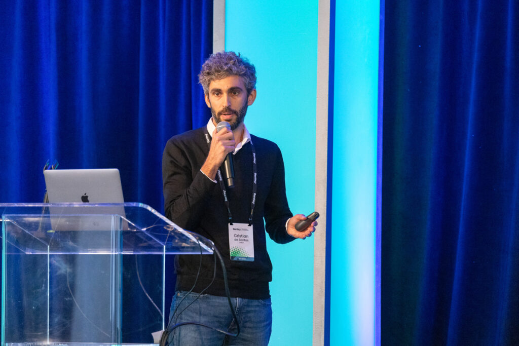 A man with curly hair presents at a conference, standing at a podium with a microphone and a remote in hand, next to a digital screen displaying a blue graphic.