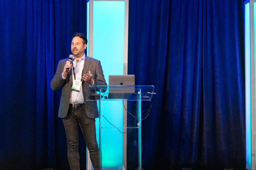 A man presenting at a conference, standing at a podium with a microphone, with a large screen beside him in a room with blue lighting.