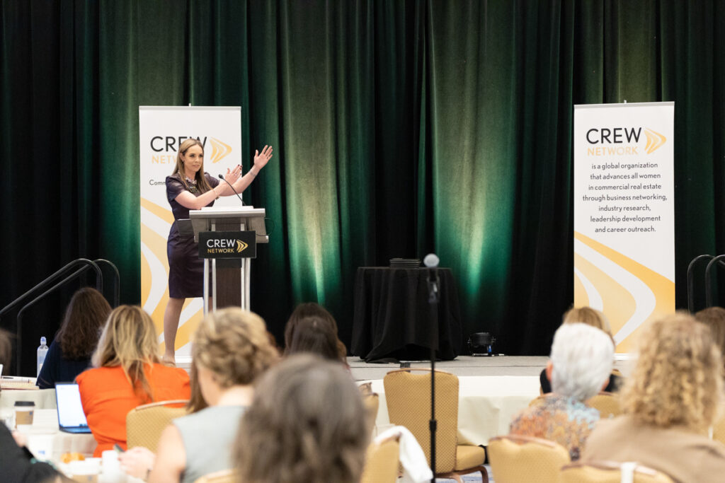 A woman gestures while giving a presentation at a podium with a crew network banner beside her to an attentive audience in a conference room.