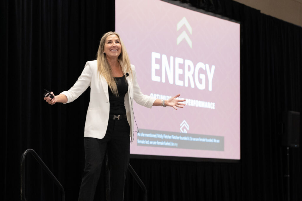 A confident woman presenting at a conference, with a clicker in one hand, standing in front of a screen displaying the word "energy" and bullet points related to attitude and performance.