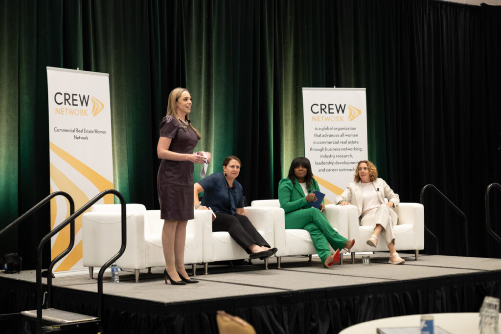 A woman stands speaking at a podium during a panel discussion at a crew network event. three other women, seated on stage, listen attentively. the setting includes banners with conference branding.