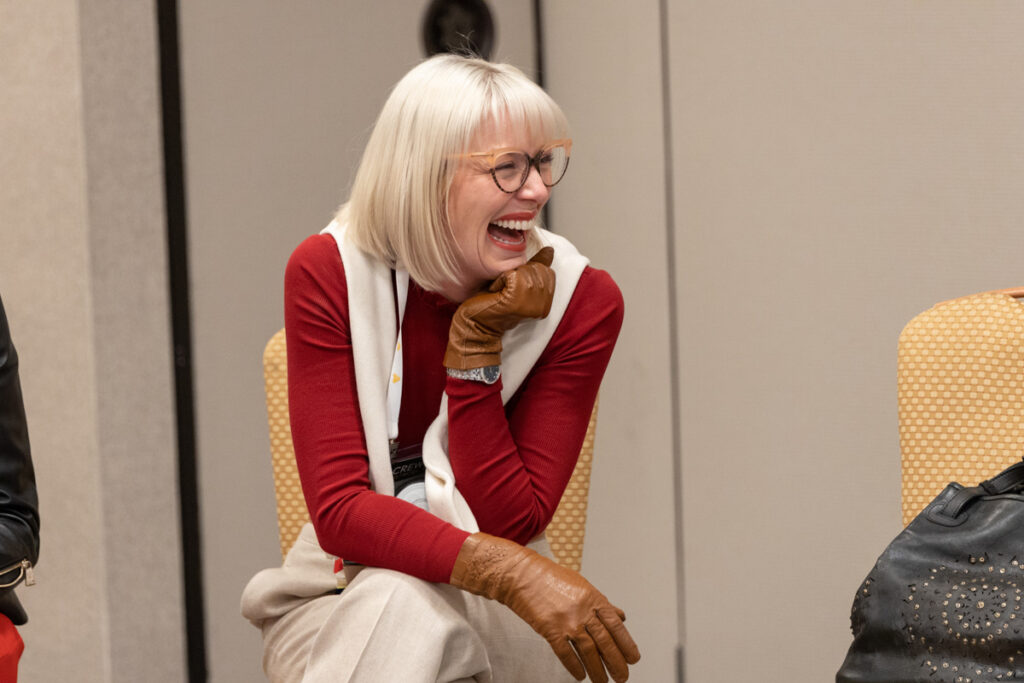 A joyful elderly woman with short white hair is laughing while wearing a white top, red cardigan, and light brown gloves, sitting in a conference room.