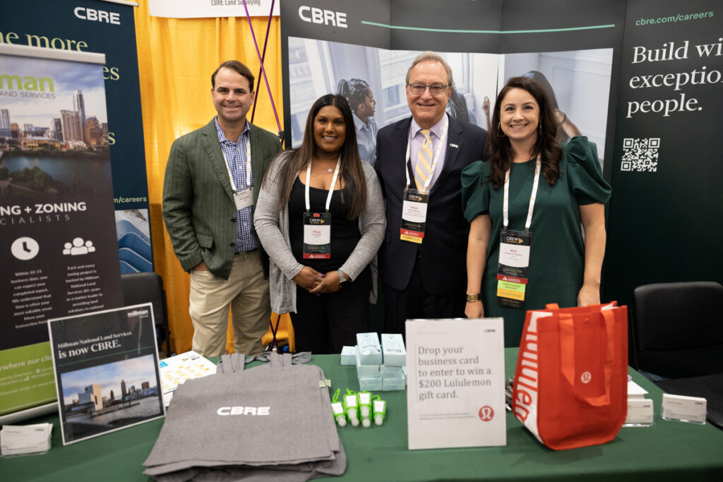 Four professionals smiling at a trade show booth labeled "cbre" with promotional items and a sign advertising a gift card giveaway.