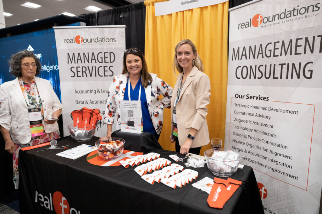 Three women stand behind a promotional booth labeled "real foundations" at a conference, smiling towards the camera. the booth displays brochures, business cards, and giveaways like foam fingers and candies.