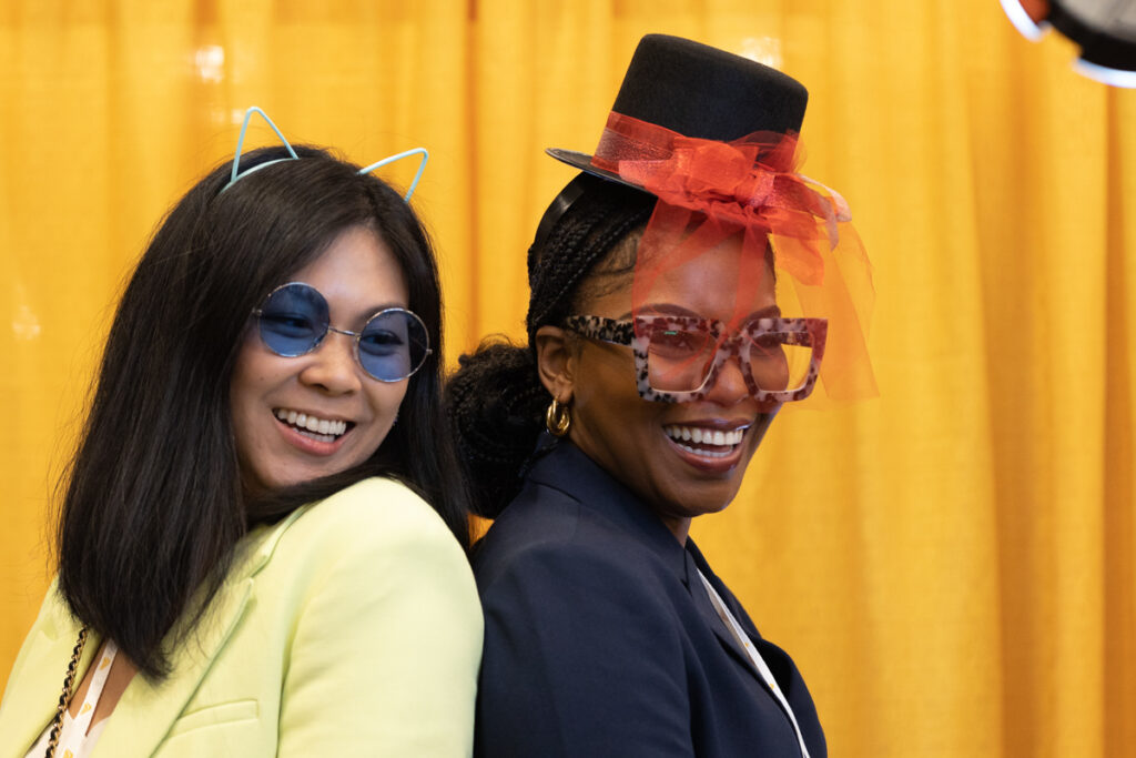 Two women smiling at a festive event, one wearing a yellow blazer and blue sunglasses, the other in a black hat with a red veil and oversized glasses. they both have decorative accessories.