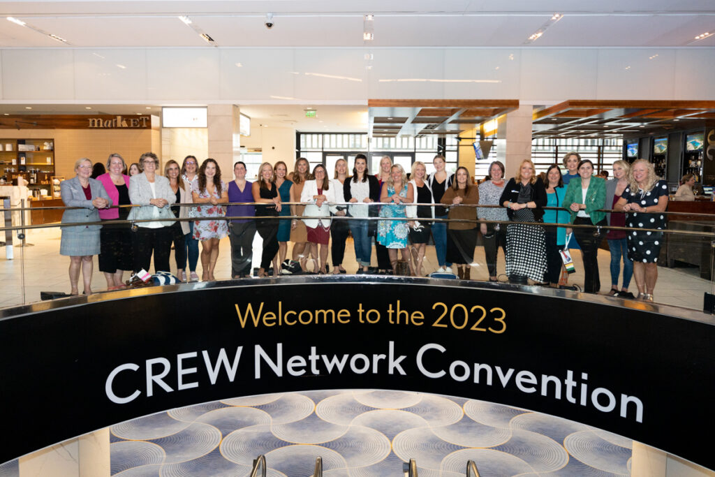 A large group of women standing behind a sign that reads "welcome to the 2023 crew network convention" in a spacious lobby.