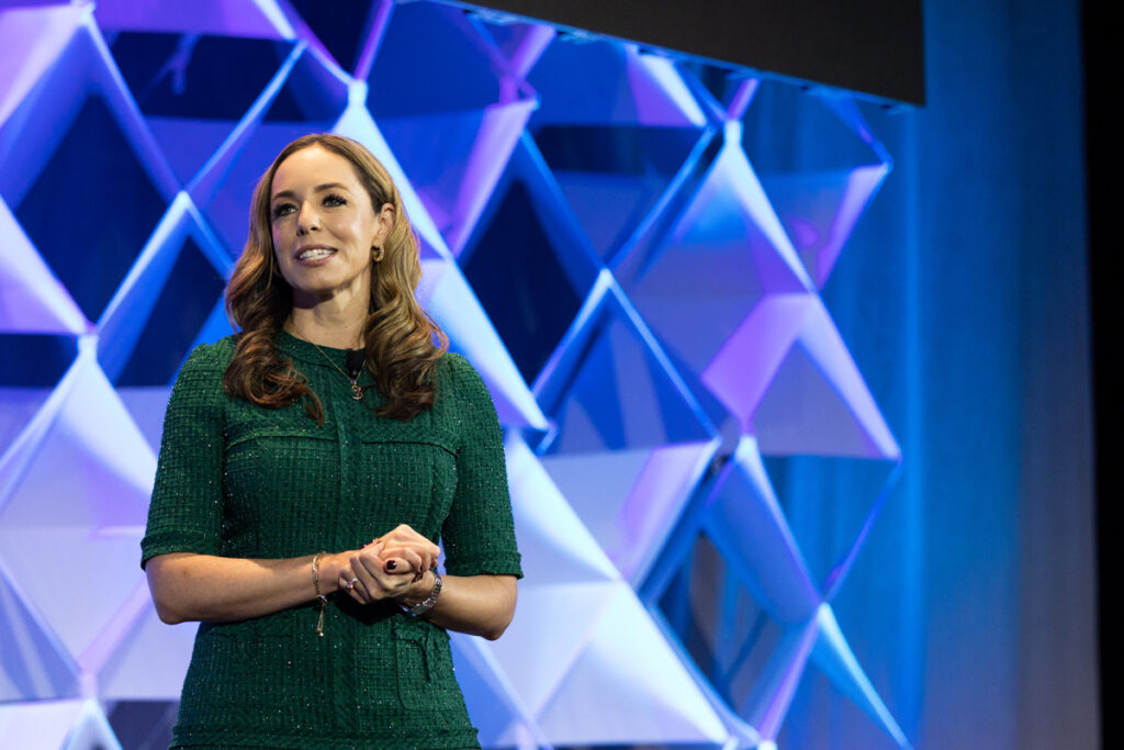 A woman in a green dress stands speaking on a stage, with a geometric blue-lit background giving a presentation. she appears confident and is gesturing slightly with her hands.