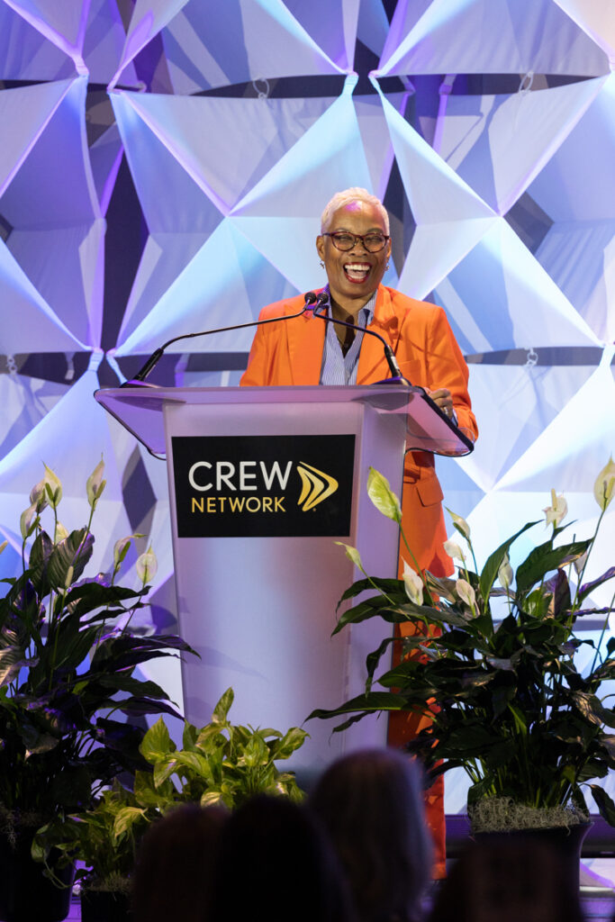 A woman in an orange jacket and glasses smiles while speaking at a podium labeled "crew network." the background features a geometric blue design.