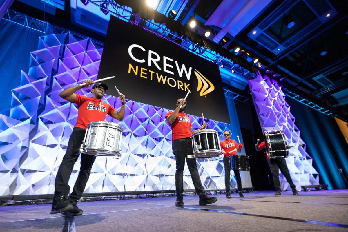 Three drummers in red shirts perform energetically on stage at a crew network event, with a large logo in the background and an illuminated geometric design.