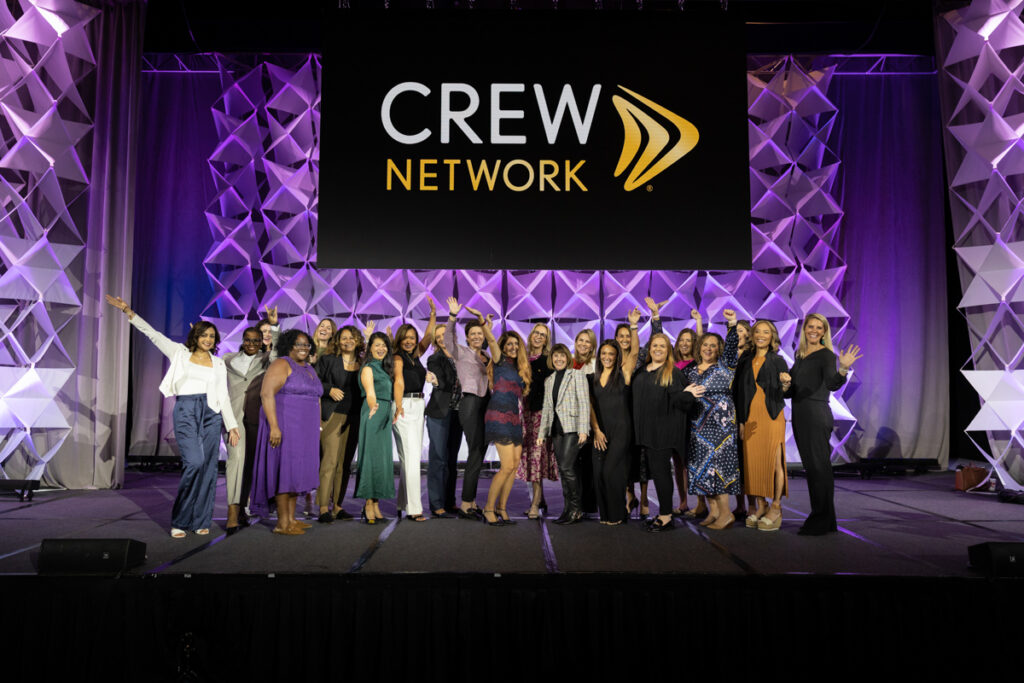 A group of diverse women cheerfully posing at a crew network event, standing on a stage with a dynamic geometric backdrop and the organization's logo prominently displayed.