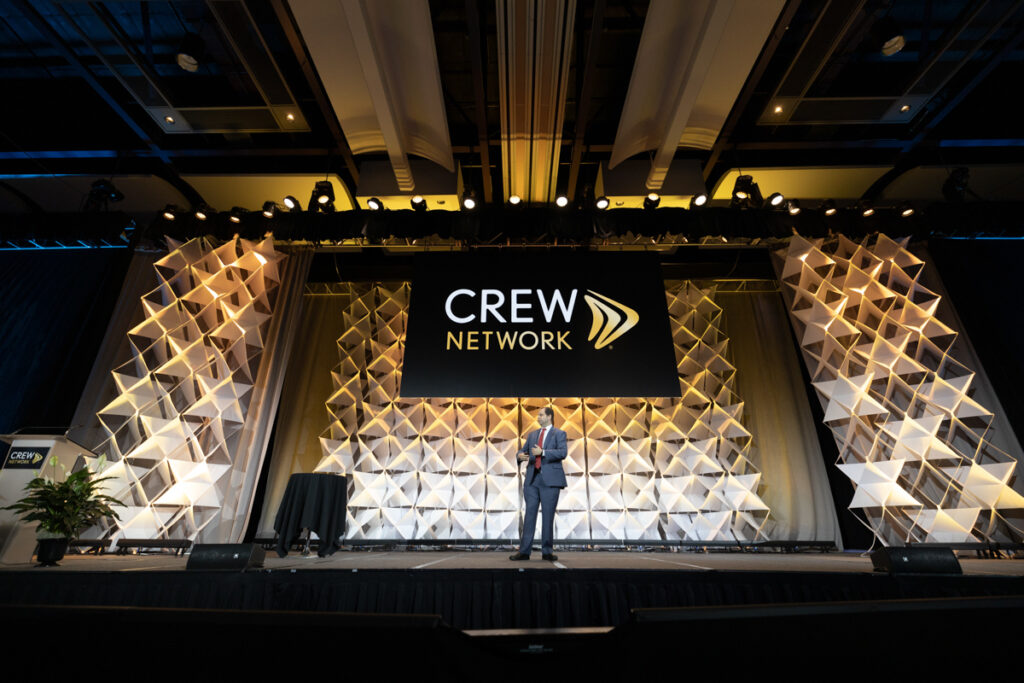 A person stands on stage at a crew network conference, presenting before a large audience with a geometrically patterned backdrop displaying the crew network logo.