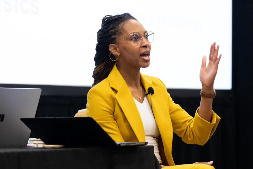 A black woman in a yellow blazer and glasses gestures passionately during a presentation at a conference, with a laptop and projector screen displaying text in the background.