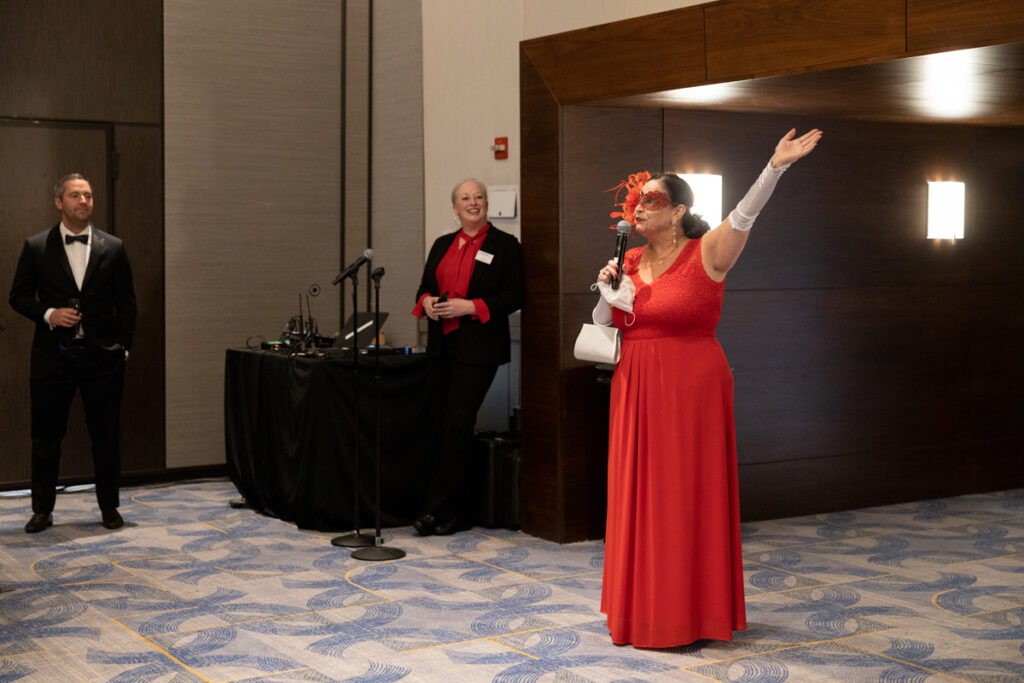 A woman in a red dress speaks animatedly at a formal event, holding a microphone, with two attentive staff members in the background.