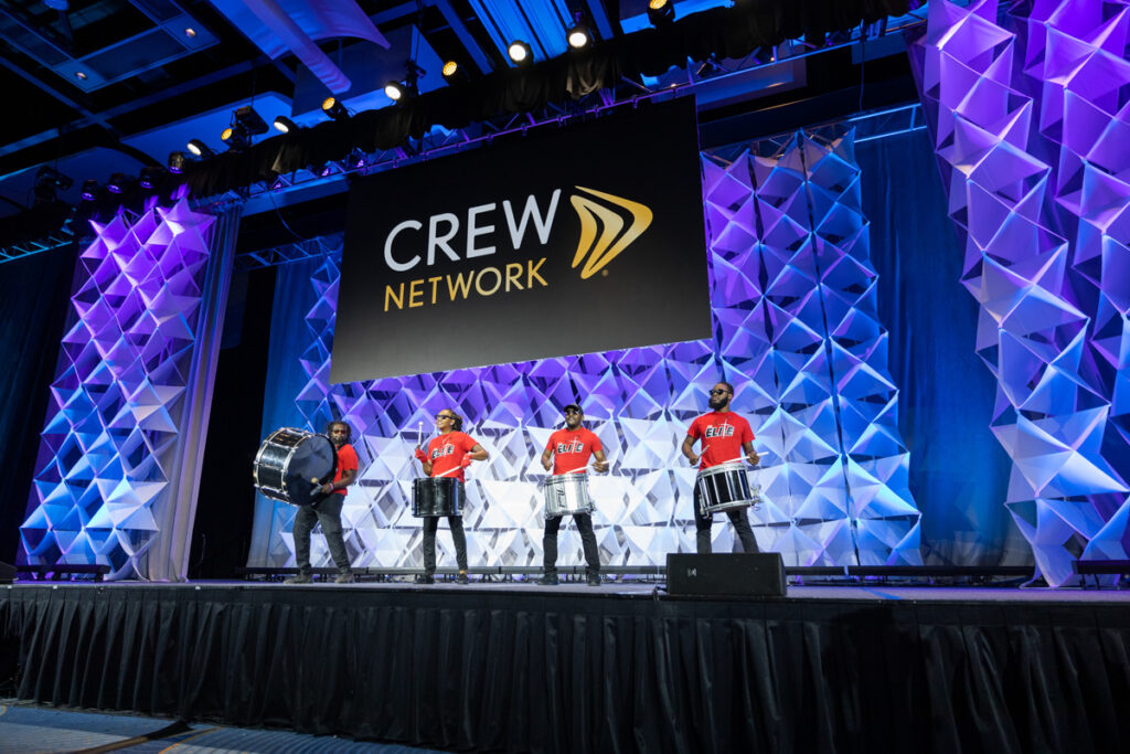 Four drummers perform on a stage at a crew network event, standing in front of a large, lit-up geometric backdrop with a prominent network logo above them.