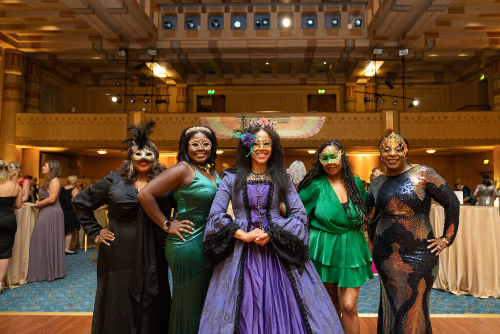 Five women dressed in elegant, colorful gowns and masquerade masks pose smiling at a festive indoor event in a grand hall.