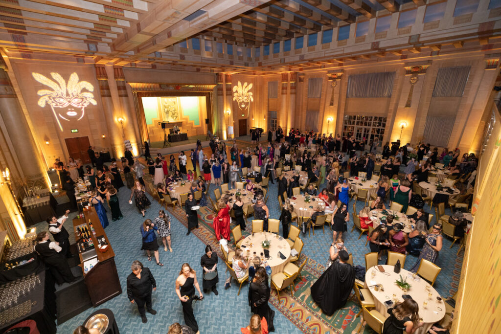 An elegant gala event in a grand ballroom with people in formal attire, sitting at round tables and mingling, with a stage and ornate decorations in the background.