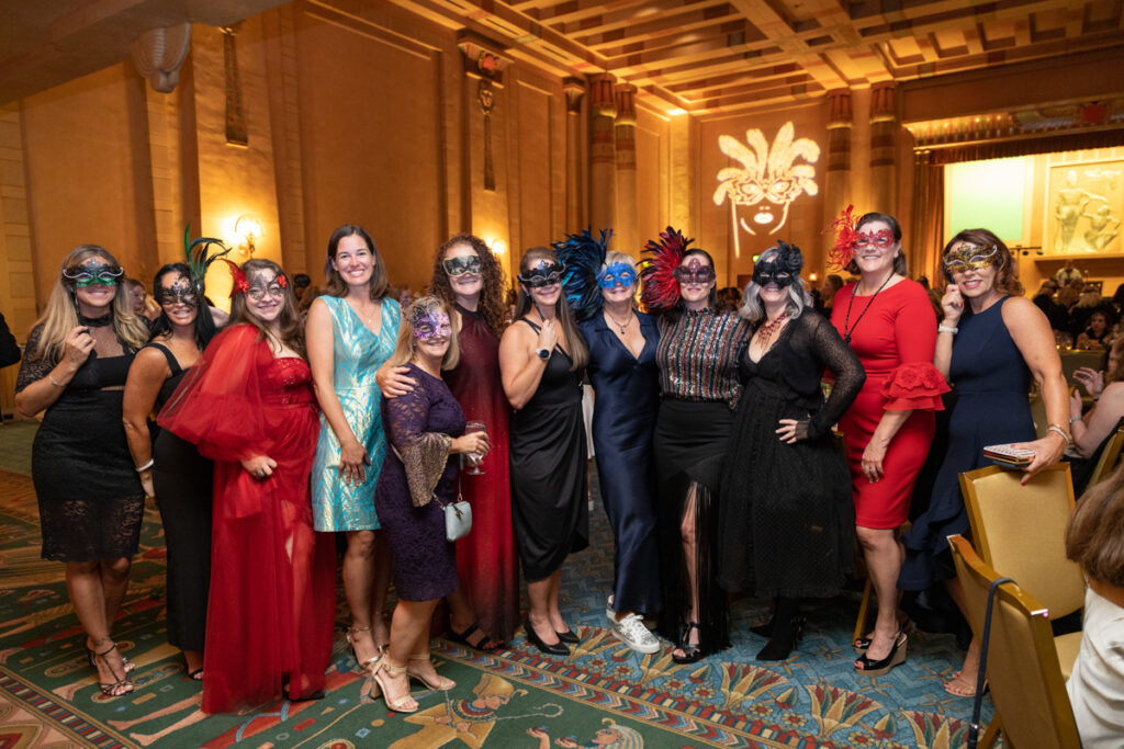 A group of women in elegant attire and colorful masks gathered at a masquerade ball in a grand, ornately decorated room.