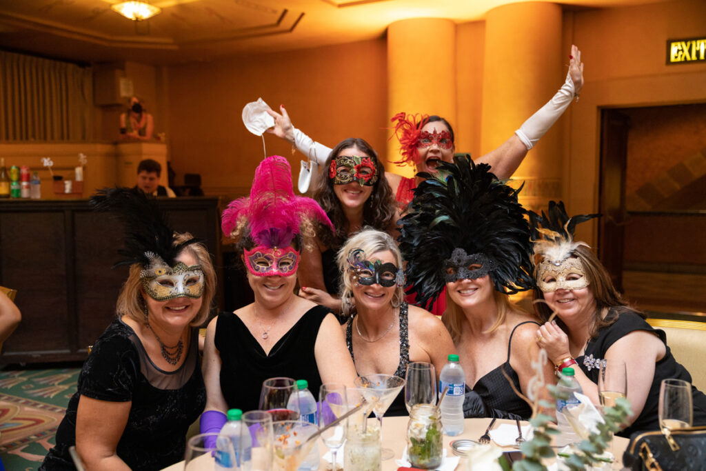 A group of seven women at a masquerade-themed party, wearing colorful masks and costumes, sitting at a table and joyfully posing for a photo.