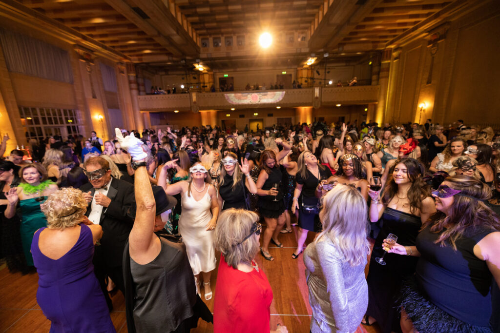 A lively masquerade ball in a large, ornate hall with guests wearing masks and elegant attire, dancing and socializing. warm lighting enhances the festive atmosphere.