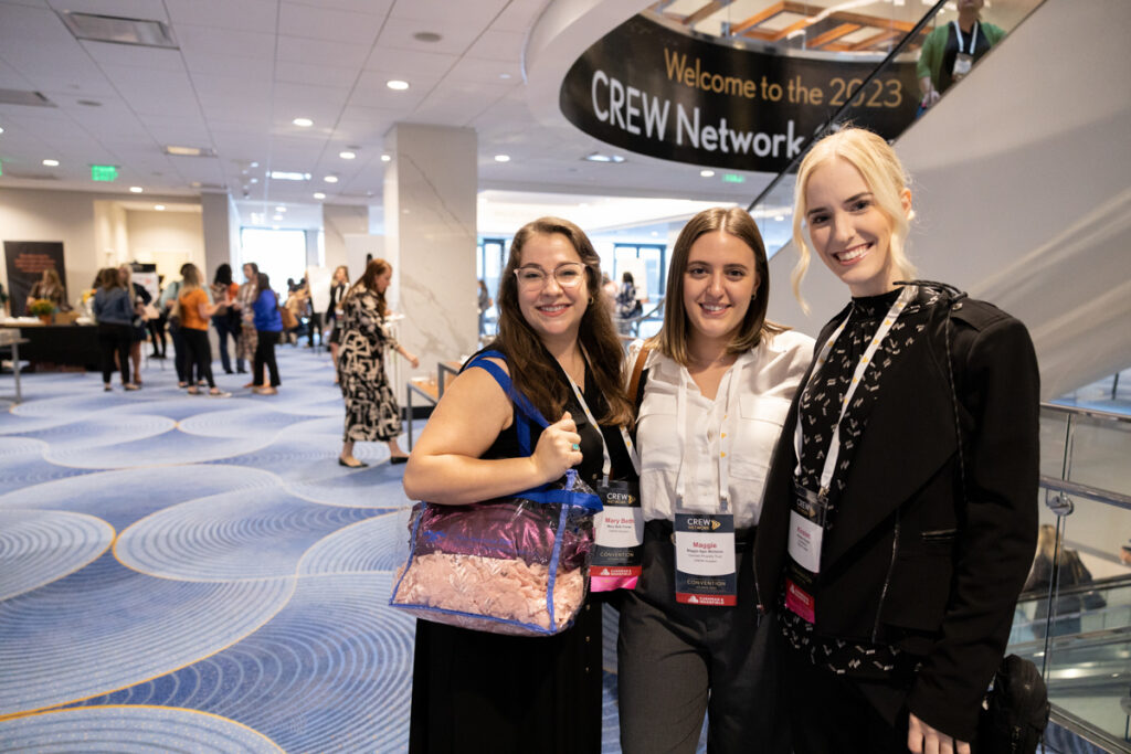 Three women smiling at a conference, holding id badges, standing in a hallway with a "welcome to the crew network" sign in the background.