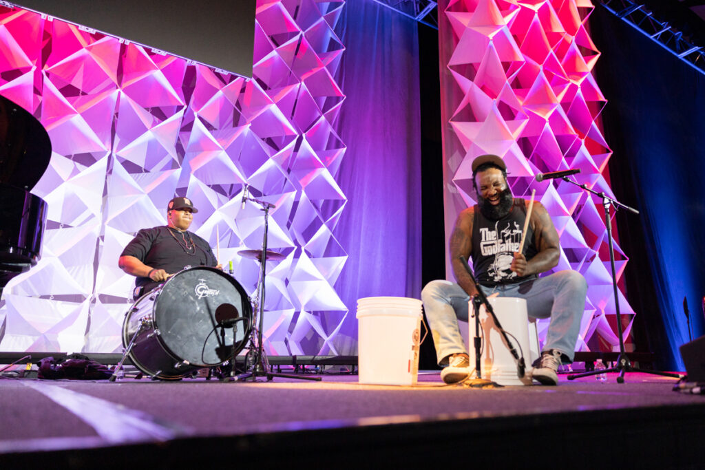 Two musicians perform on stage at a conference, one playing a drum kit and the other on a cajón, against a brightly lit geometric backdrop.