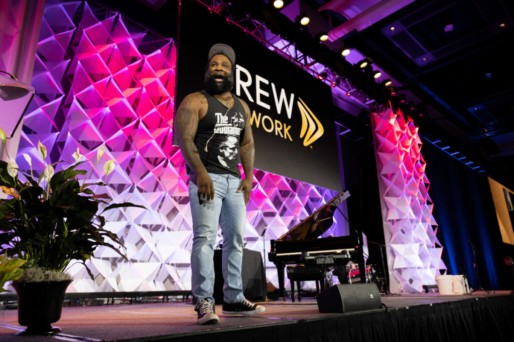 A man with a beard, wearing a tank top, jeans, and a beanie, smiles and walks on stage at an event hall with colorful geometric decorations and the text "brew network" in the background.