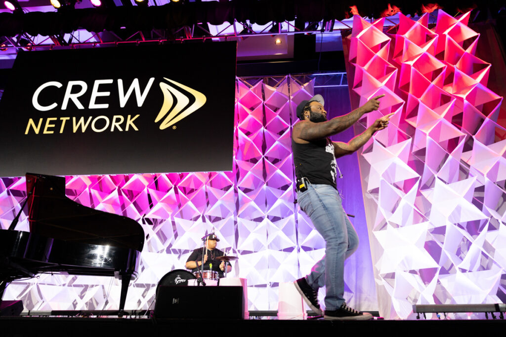 A man energetically singing on stage at a crew network event, with a drummer in the background, against a backdrop of pink lit geometric patterns.