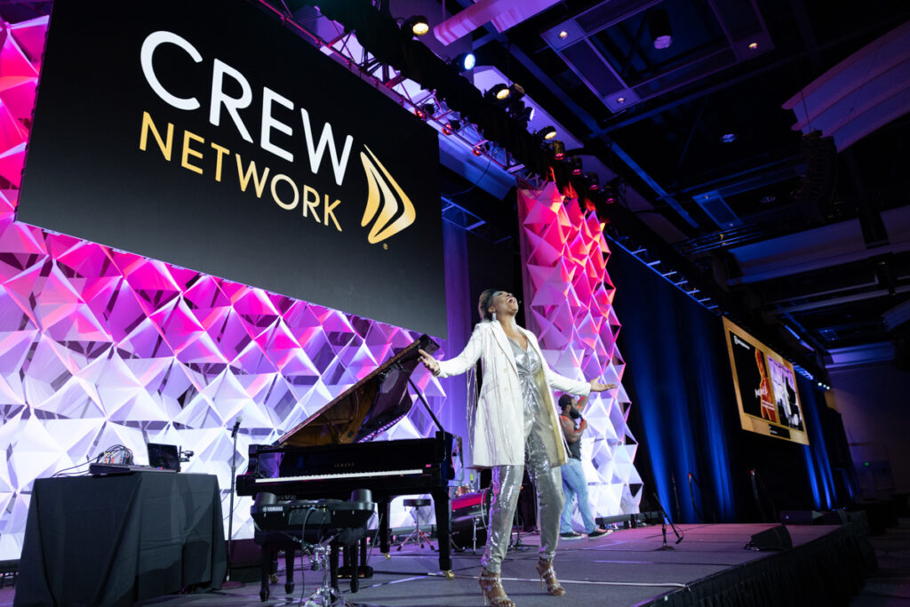 A woman performs on stage beside a grand piano at a crew network event, with a large, colorful geometric backdrop and the organization's logo displayed prominently.