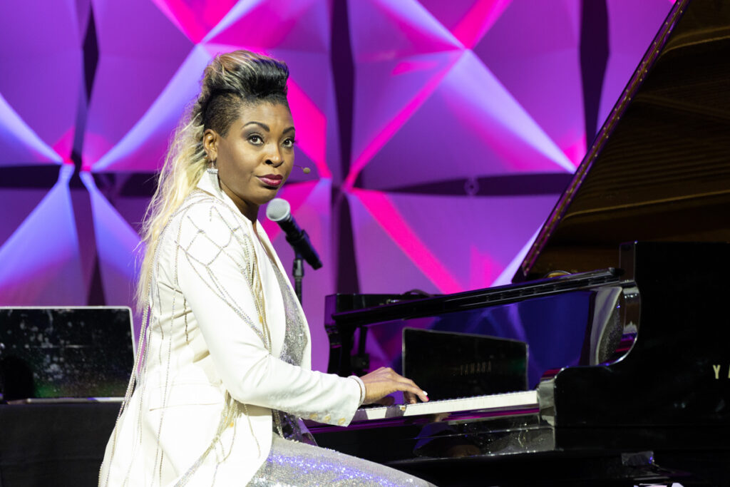 A woman in a glittering white dress plays a grand piano on a stage with purple geometric backdrops, looking over her shoulder with a slight smile.