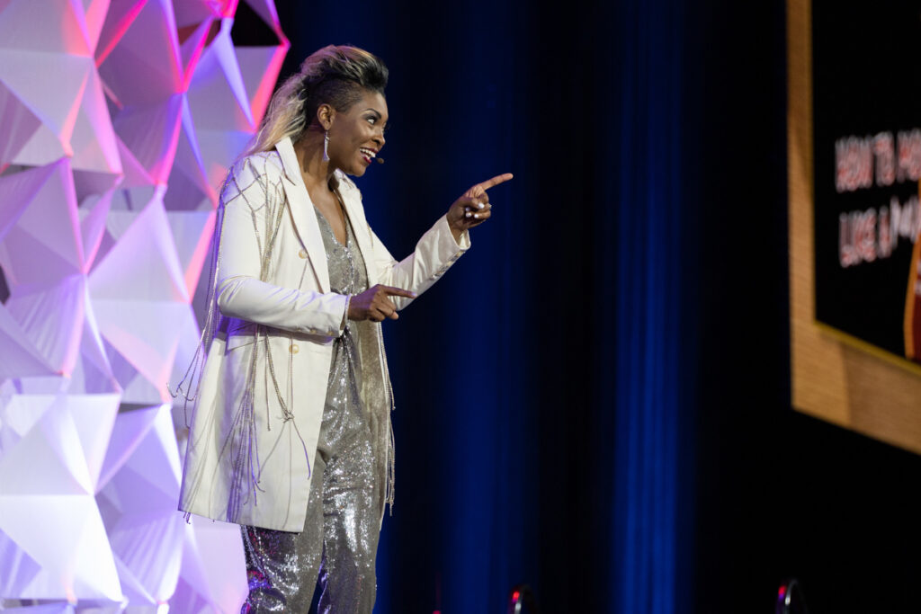 A dynamic speaker on stage pointing into the audience, dressed in a sparkling white and gold outfit, with a large graphic screen in the background.