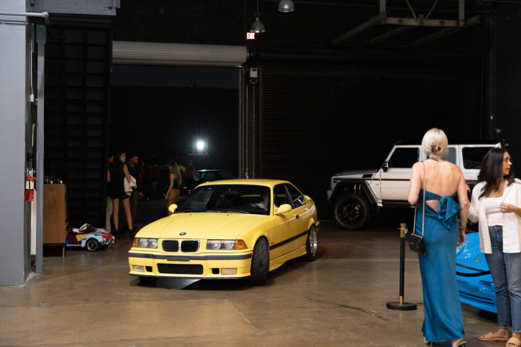 A yellow bmw sports car parked on display in a warehouse setting with open garage doors and a group of people standing in the background.