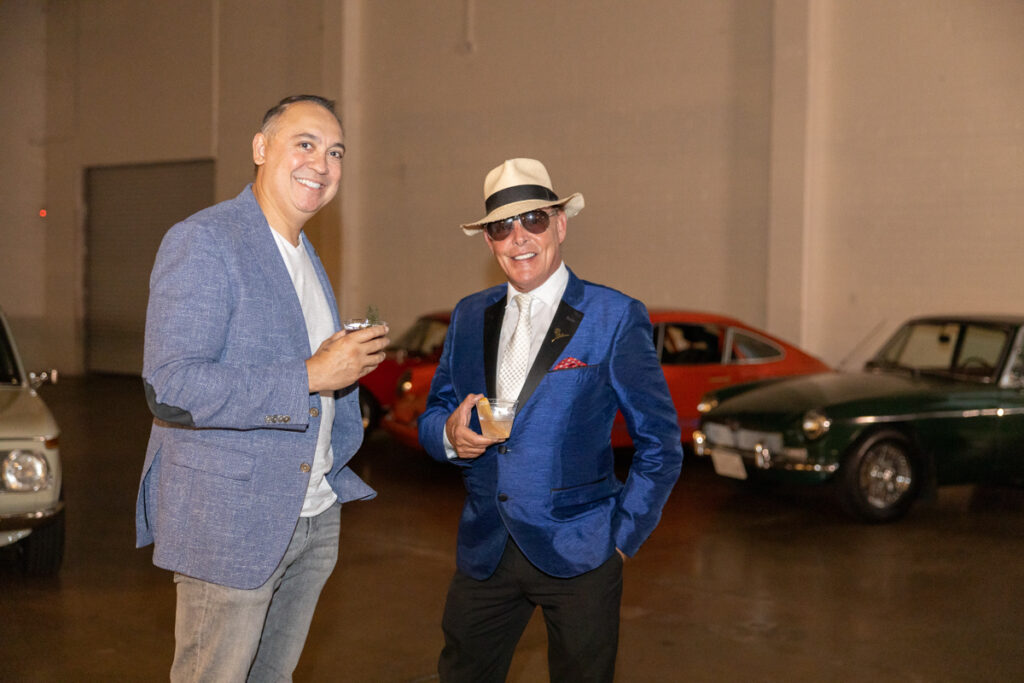 Two smiling men in stylish suits, one in blue and the other in gray, stand in a garage with vintage cars in the background. the man in blue is holding a smartphone.