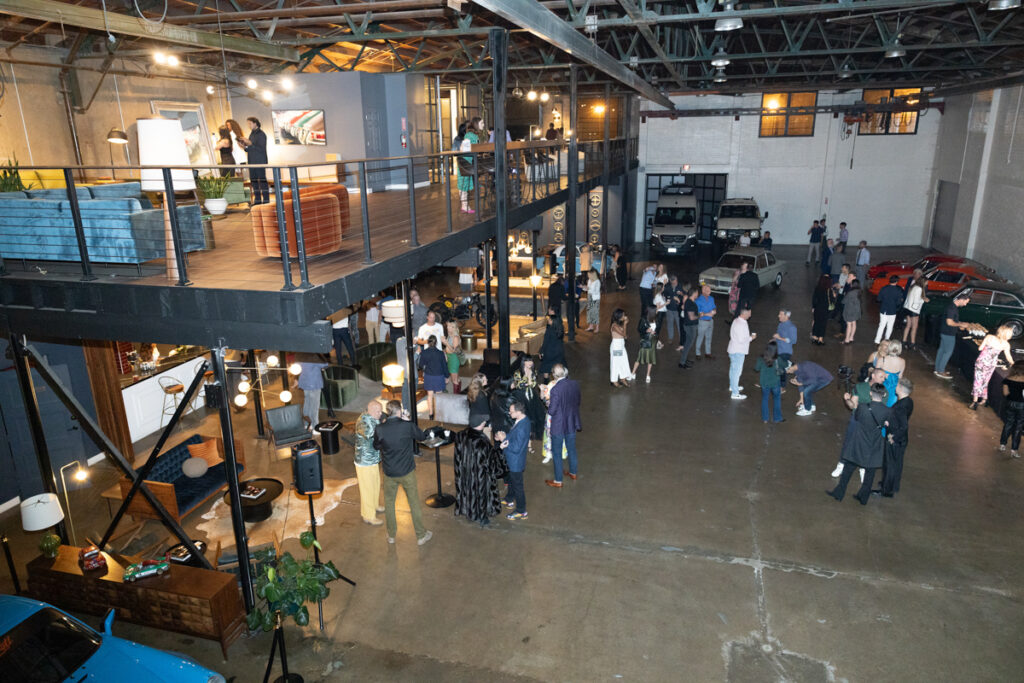 A bustling indoor event in a spacious industrial-style venue with exposed beams and a mezzanine. people mingle near display stalls and vintage cars.