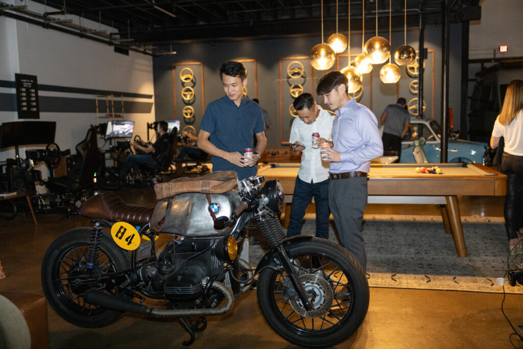 Three men examining a vintage motorcycle in a trendy indoor space with decorative light bulbs and a bar in the background, conveying a casual social atmosphere.