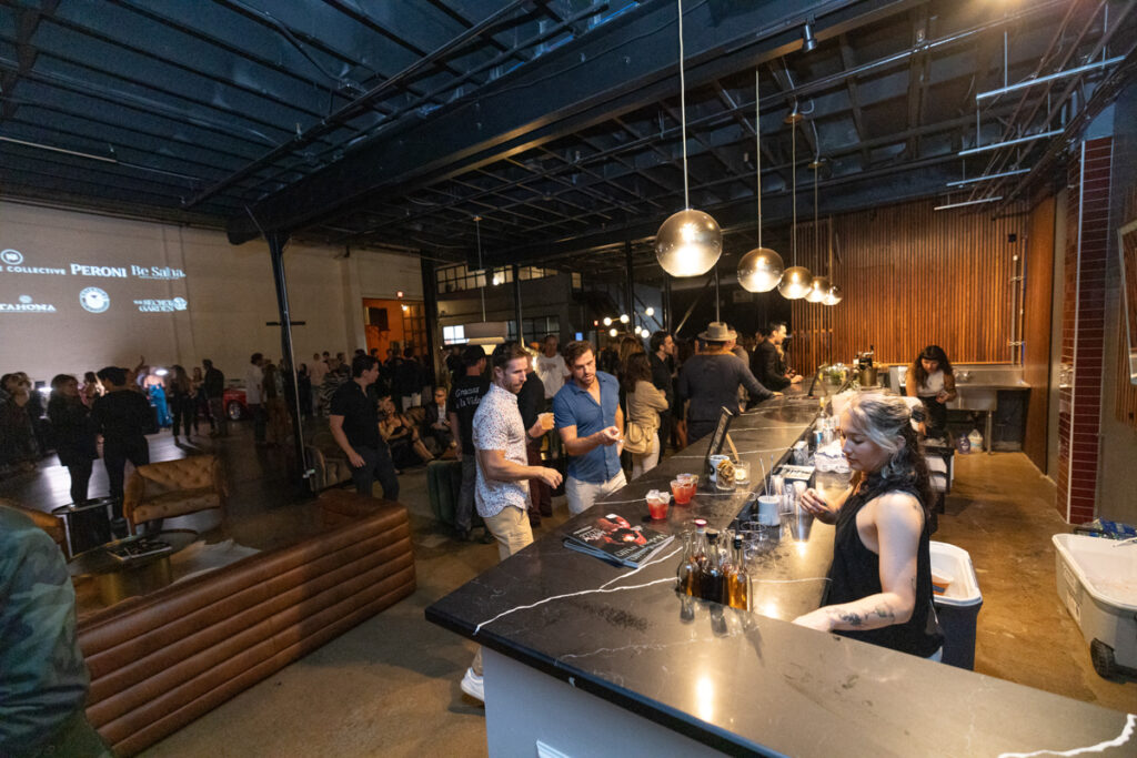 A bustling indoor event at a bar with people socializing and bartenders serving drinks. the setting features industrial decor with hanging lamps and a projector screen displaying text.