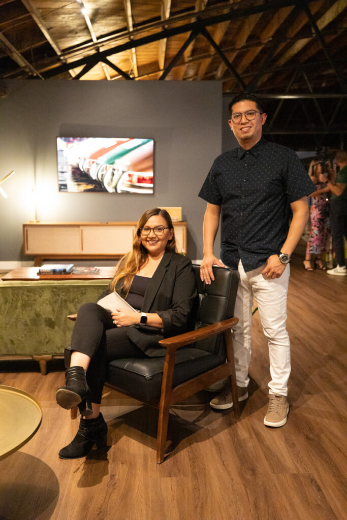 A man standing and a woman sitting in a stylishly furnished room with wood ceilings and modern decor, both smiling at the camera.