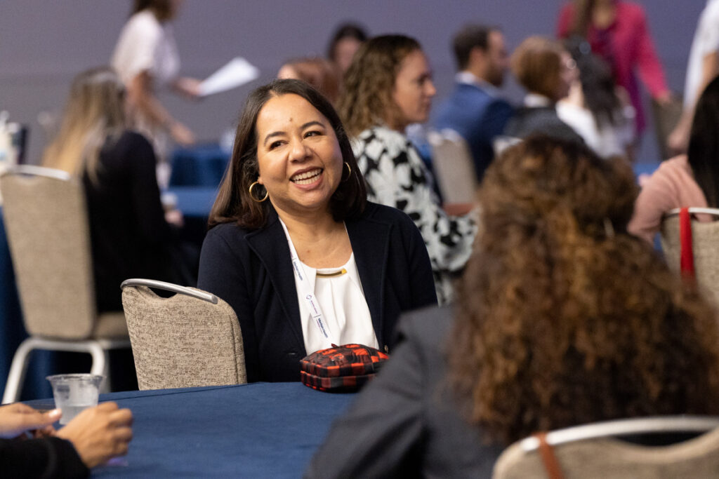 A smiling woman engaging in conversation at a conference table, surrounded by other attendees in a bustling hall. she wears a white blouse and a necklace, exuding a friendly demeanor.