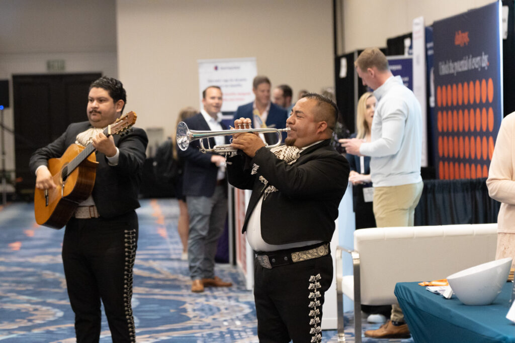 Two mariachi musicians performing at a business event, one playing a guitar and the other a trumpet, with attendees standing in the background.