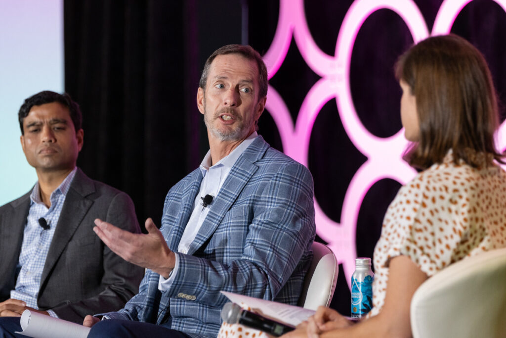 Panel discussion at a conference with a man speaking animatedly, flanked by a focused male listener on the left and a female listener on the right, all sitting against a backdrop with a pink logo.