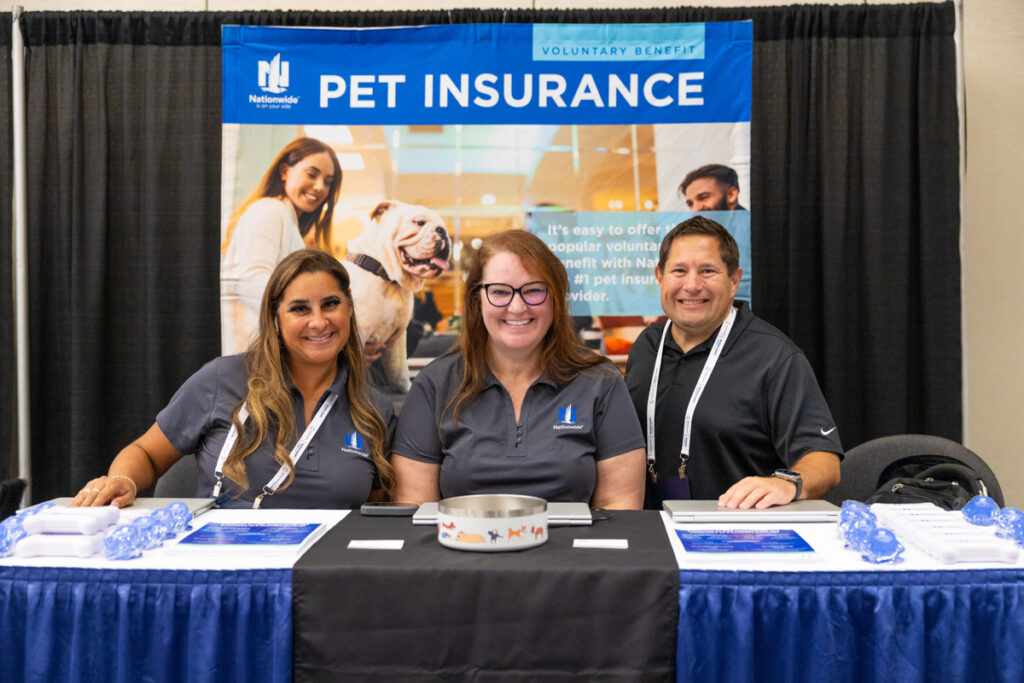 Three smiling people at a "nationwide pet insurance" promotional booth, with pamphlets and promotional items displayed on the table in front of them.