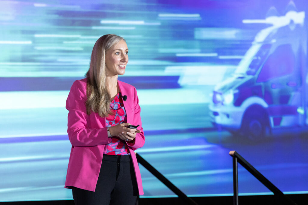 A woman wearing a pink blazer presenting at a tech conference with a background image of a fast-moving ambulance. she is holding a remote and smiling while speaking.