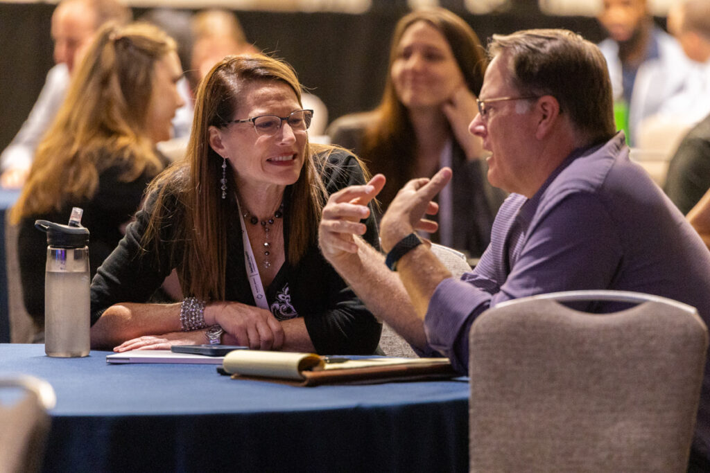 A woman and a man engage in a lively conversation across a table at a conference, surrounded by other attendees.