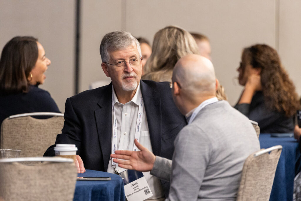 An older man with glasses in a meeting, actively engaged in conversation with two other attendees at a conference table. he wears a dark suit and a name badge.