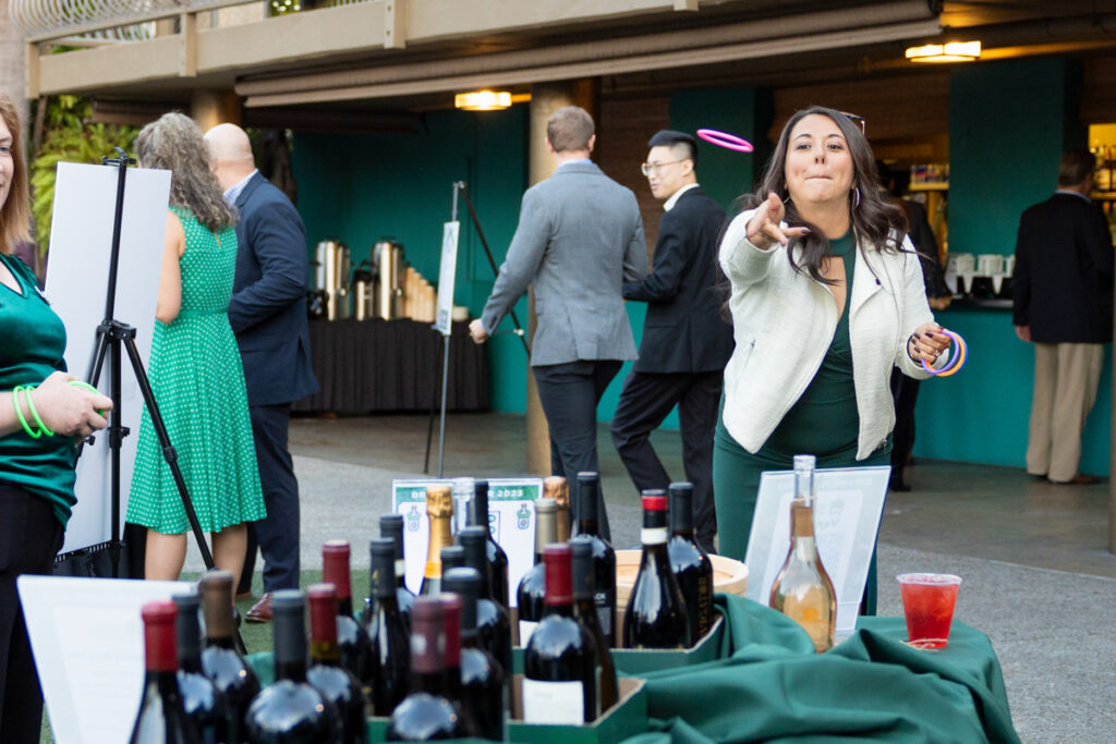 A woman at an outdoor networking event playfully throws a small object, smiling towards the camera, as people mingle and browse items on tables with wine bottles.