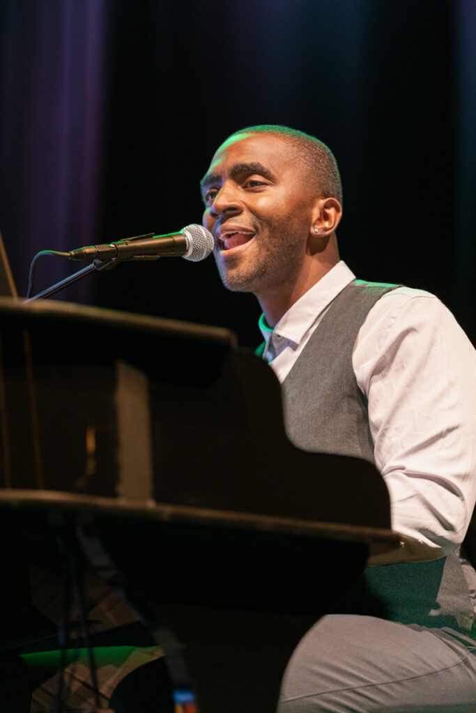 A man smiling and singing into a microphone while playing the piano on a stage, wearing a gray vest and white shirt.