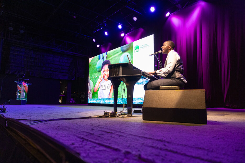 A man presenting on stage at a conference, standing by a podium with a large screen behind him showing a smiling woman's face, illuminated by stage lighting with a purple backdrop.