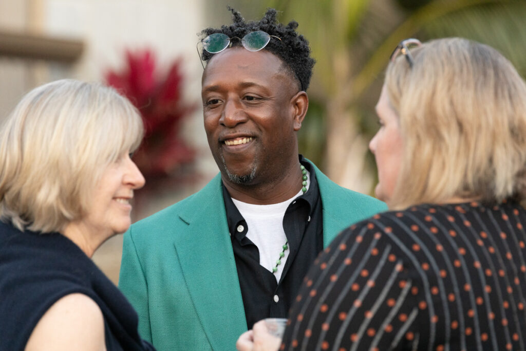 A man in a green blazer smiles while conversing with two women at an outdoor social event, holding a glass in his hand.