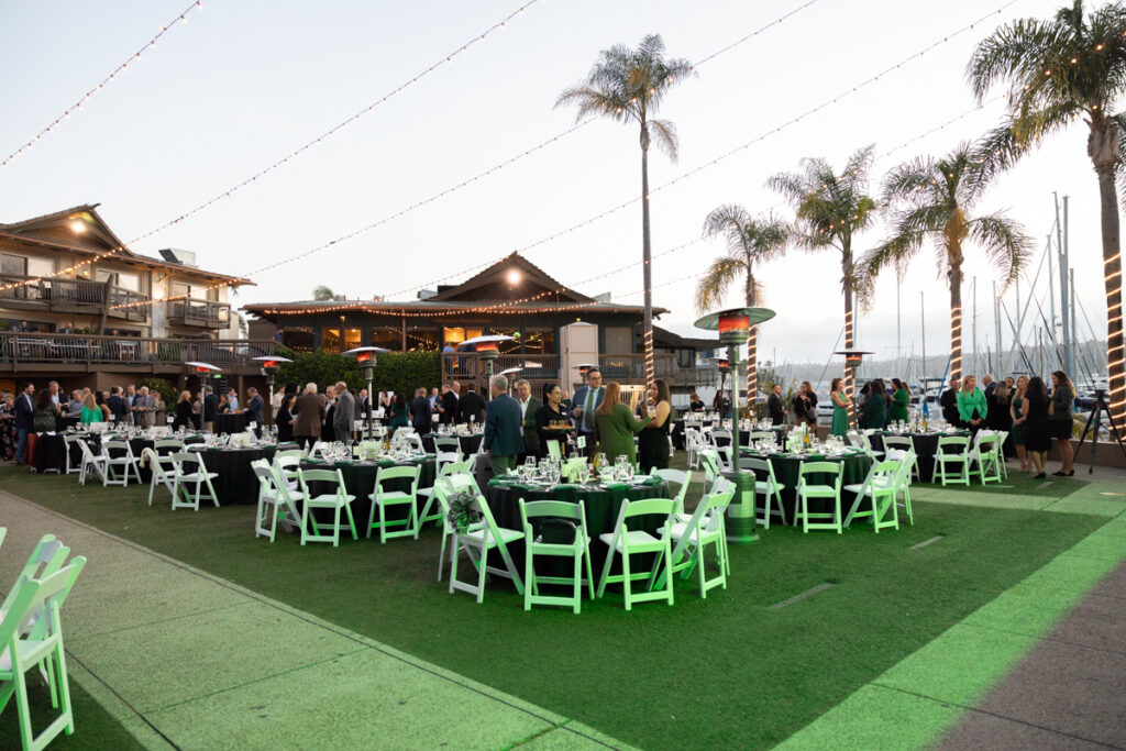 Outdoor event setup with tables covered with white linens, guests mingling, and festoon lights overhead, beside a waterfront and palm trees at dusk.